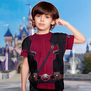 May include: A red and black t-shirt featuring a Deadpool costume design. The shirt has a detailed print of the character's suit, including a belt with a Deadpool logo. The short-sleeved shirt is worn by a child.