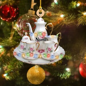 Peut inclure: Ornement de Noël en porcelaine blanche représentant un service à thé. L'ensemble comprend une théière, un sucrier, une crémière, deux tasses et soucoupes, et un plateau. L'ensemble est décoré de roses roses et de bordures dorées. Le mot "Name" est imprimé sur la théière.