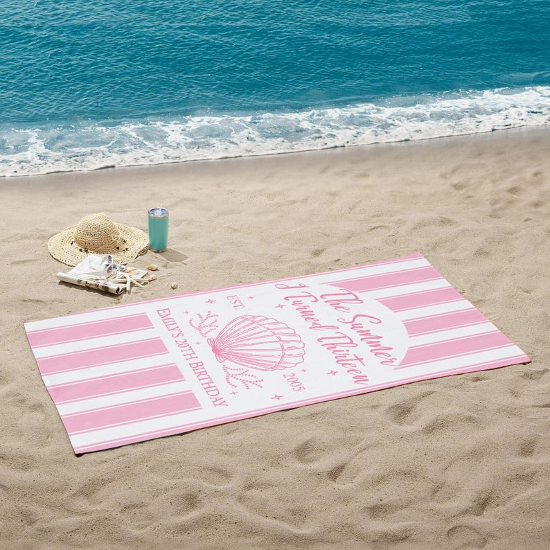 May include: A pink and white striped beach towel with the text "The Summer I Turned Thirteen" and "Emily's 20th Birthday" printed on it. A straw hat, a turquoise tumbler, and seashells are on the sand.