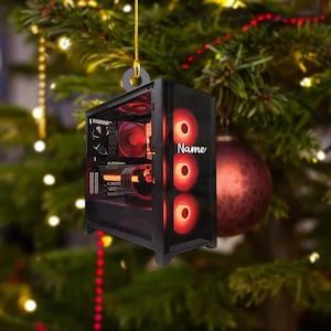 May include: A black computer case Christmas ornament with red interior lighting and the word "Name" in white. The ornament is hanging from a gold string against a backdrop of a Christmas tree with ornaments.