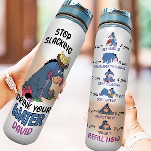 Peut inclure: Une bouteille d'eau blanche avec un couvercle bleu et un personnage de dessin animé bleu et violet d'Eeyore de Winnie l'ourson. La bouteille porte le texte "Stop Slacking" et "Drink Your Water David". La bouteille comporte également un horaire avec des images d'Eeyore pour chaque heure.
