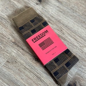 Op de afbeelding: Een paar bruine sokken met een zwart-wit Amerikaans vlagpatroon. De sokken zijn verpakt in een rode doos met de woorden "FREEDOM SOCKS" en "Designed in the USA". De doos vermeldt ook "Maat: L".