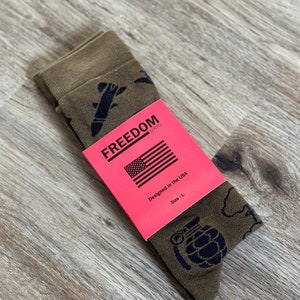 May include: A pair of brown socks with a black print of military-themed graphics, including a grenade, a plane, and a bomb. The socks are folded and packaged with a red label that says "FREEDOM SOCKS" and "Designed in the USA". The label also indicates the size is "L".