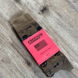 May include: A pair of brown socks with a black alien and UFO print. The socks are folded and have a red tag that says "FREEDOM socks" with an American flag and the text "Designed in the USA" and "Size: L".