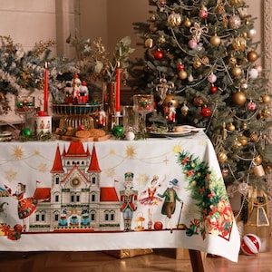 Puede incluir: Un mantel blanco con un diseño de Cascanueces que presenta un castillo, una bailarina, un soldado cascanueces y un ratón. El mantel está puesto para una cena de Navidad con un árbol de Navidad decorado en el fondo.