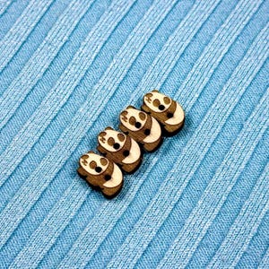 Puede incluir: Cuatro botones de madera con forma de panda, con detalles en blanco y negro, dispuestos en diagonal sobre un fondo de tela acanalada azul claro. Los botones tienen un acabado de madera natural y la tela tiene un aspecto texturizado.