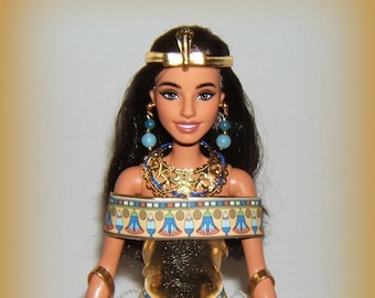 May include: A doll dressed as an Egyptian queen, wearing a gold and cream-colored gown with intricate detailing. The doll has dark hair, blue earrings, and a gold headdress. The gown features a white cape.