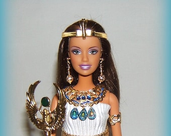 May include: A doll dressed in an Egyptian-themed outfit. The doll wears a white dress with gold accents, a gold headpiece, and matching jewelry. She holds a gold staff with an ornate design and a green gem.