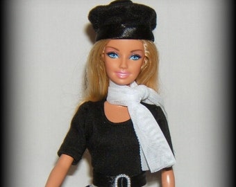May include: A black and white gingham skirt with a black top and a white scarf. The doll is wearing a black beret and has blue eyes and blonde hair. She is holding a black handbag.