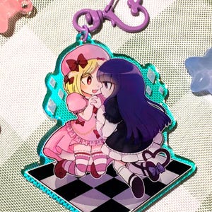 Könnte beinhalten: Ein klarer Acryl-Anhänger mit zwei Mädchen im Anime-Stil, eine blonde in Rosa und die andere mit dunklem Haar in Schwarz und Weiß. Der Anhänger hat einen türkisfarbenen Rand und eine karierte Basis. Sternförmige Anhänger in Rosa, Lila und Blau umgeben den Hauptanhänger.