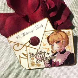 May include: An enamel pin shaped like an envelope, featuring the text "To. Ushiromiya Family" and "Welcome to Rokkenjima". The pin showcases a cartoon illustration of a woman with blonde hair and a black dress. A burgundy rose is in the background.