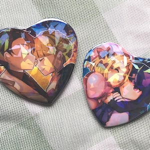 Puede incluir: Dos pines en forma de corazón con ilustraciones de estilo anime de parejas abrazándose. Los pines tienen un acabado brillante y un fondo geométrico colorido. Un pin presenta una pareja de pelo castaño, el otro una pareja rubia y morena.