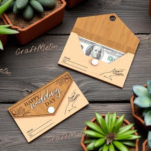 K&ouml;nnte beinhalten: Zwei Geldhalter aus Holz, einer mit einer sichtbaren Hundert-Dollar-Note und der andere mit dem Text "Happy Wedding Day". Beide haben ein Handdesign und einen Knopfverschluss. Die Halter liegen auf einer dunklen Holzoberfl&auml;che, umgeben von Topf-Sukkulenten.