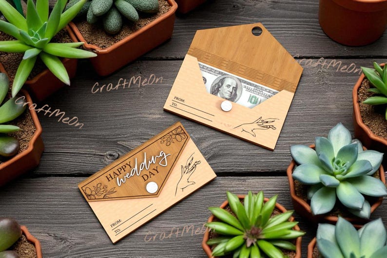 K&ouml;nnte beinhalten: Zwei Geldhalter aus Holz, einer mit einer sichtbaren Hundert-Dollar-Note und der andere mit dem Text "Happy Wedding Day". Beide haben ein Handdesign und einen Knopfverschluss. Die Halter liegen auf einer dunklen Holzoberfl&auml;che, umgeben von Topf-Sukkulenten.
