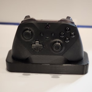May include: A black Xbox controller is sitting on a black charging stand. The controller has a black and white design with a cross in the center.