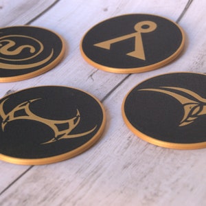 May include: Four round black coasters with gold trim and different symbols. The symbols are a crescent moon with a letter S inside, a stylized letter A with a circle on top, a stylized bird with wings spread, and a stylized three-pointed star.