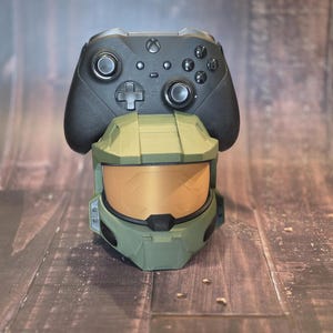 May include: A black Xbox controller rests atop a green and gold helmet, likely a gaming accessory. The helmet has a visor and is designed to hold the controller. The items are on a wooden surface.