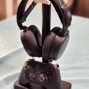 May include: A black gaming headset with a green and black patterned headband is displayed on a black stand with a black Xbox controller.