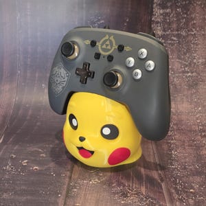 May include: A gray wireless video game controller featuring a gold Triforce emblem and a Hylian crest, resting on a yellow Pikachu-shaped stand. The controller has black analog sticks, a black directional pad, and silver A, B, X, and Y buttons.
