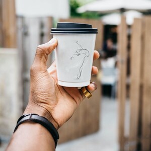 Può includere: Un bicchiere di caffè da asporto bianco con coperchio nero. Il bicchiere ha un disegno a linea di una persona che balla.