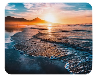 Pode incluir: Um pôr do sol cênico sobre um oceano calmo com ondas quebrando suavemente na costa. O céu é uma mistura vibrante de laranja, rosa e azul, com nuvens fofas. O sol se põe atrás de uma cordilheira ao longe.
