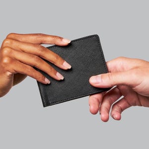 May include: A black wallet with a textured surface is being passed between two hands against a gray background. The wallet appears to be made of leather or a similar material. The image is well-lit and focuses on the exchange.