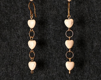 May include: A pair of dangling earrings with a gold-colored hook and chain design. Each earring features four off-white heart-shaped beads connected by small gold rings. A small pearl bead is at the top of each earring.