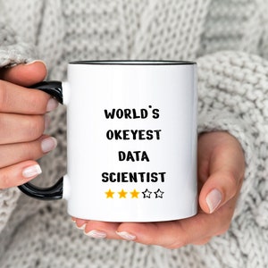 Op de afbeelding: Een witte keramische mok met een zwarte rand en handvat. De mok heeft zwarte tekst die "World's Okayest Data Scientist" luidt met vijf gele sterren onder de tekst.