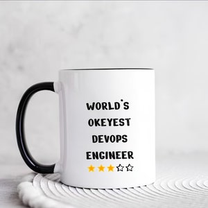 Könnte beinhalten: Weiße Keramiktasse mit schwarzem Henkel und Rand. Die Tasse hat schwarzen Text, der "WORLD'S OKEYEST DEVOPS ENGINEER" lautet, mit vier gelben Sternen unter dem Text.