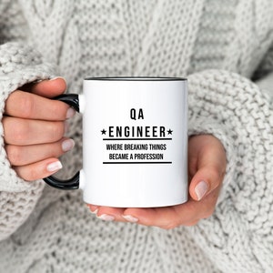 Può includere: Tazza in ceramica bianca con bordo e manico neri. La tazza ha un testo nero che recita "QA *ENGINEER* WHERE BREAKING THINGS BECAME A PROFESSION"