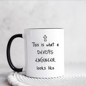 Könnte beinhalten: Eine weiße Keramiktasse mit schwarzem Henkel und schwarzem Rand. Die Tasse hat einen schwarzen Text, der "This is what a DEVOPS ENGINEER looks like" lautet, mit einem nach oben zeigenden Pfeil.