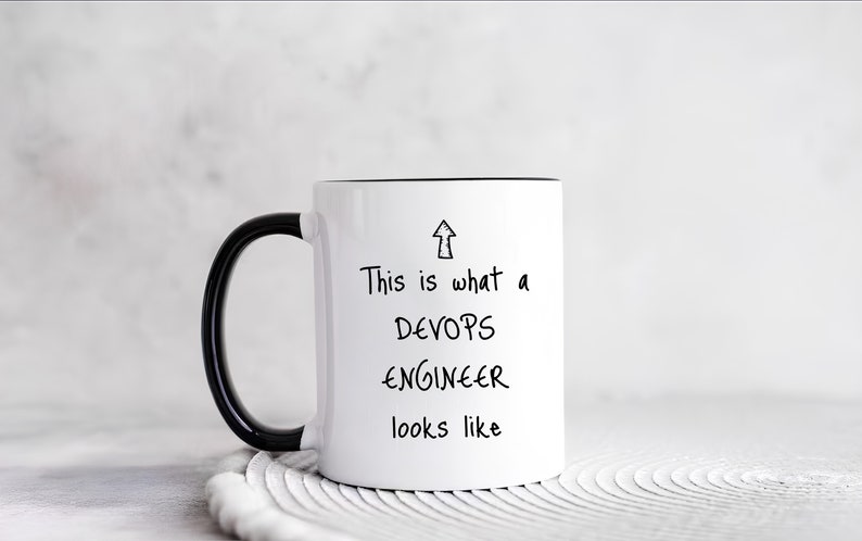 K&ouml;nnte beinhalten: Eine wei&szlig;e Keramiktasse mit schwarzem Henkel und schwarzem Rand. Die Tasse hat einen schwarzen Text, der "This is what a DEVOPS ENGINEER looks like" lautet, mit einem nach oben zeigenden Pfeil.