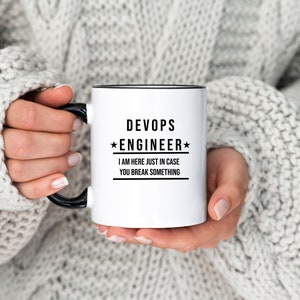 Może przedstawiać: Biały kubek ceramiczny z czarnym rantem i uchwytem. Kubek ma czarny napis: "DevOps *Engineer* I am here just in case you break something."