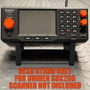 Puede incluir: Un soporte de escritorio negro para un escáner Uniden SDS200. El soporte está hecho de plástico y tiene un diseño simple. El texto "DESK STAND ONLY FOR UNIDEN SDS200 SCANNER NOT INCLUDED" está impreso en rojo sobre un fondo blanco.