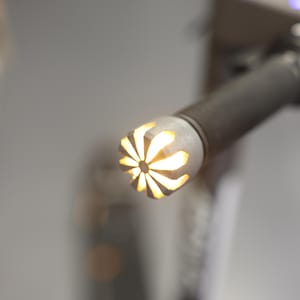 Op de afbeelding: Close-up van een zilverkleurig, cilindrisch object met een getextureerde grip. Het uiteinde heeft een bloemachtig ontwerp met geel licht dat door de uitgesneden vormen schijnt. Het object lijkt deel uit te maken van een groter apparaat.