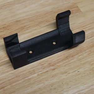 May include: A black 3D printed wall mount with two holes for mounting screws. The mount has a curved section on one end for holding a cylindrical object.