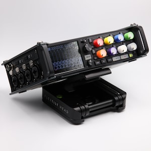 Może przedstawiać: Czarny rejestrator audio z kolorowymi pokrętłami i cyfrowym wyświetlaczem, zamontowany na czarnym statywie "FLEXTILT HEAD". Rejestrator posiada wiele gniazd wejściowych i ekran wyświetlający poziomy dźwięku. Stojak umożliwia ustawienie pod kątem.