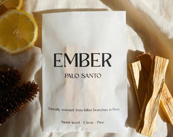 Könnte beinhalten: Ein weißer Papierbeutel mit der Aufschrift EMBER PALO SANTO und dem Text Ethically sourced from fallen branches in Peru sowie Sweet wood - Citrus - Pine. Der Beutel ist von zwei gelben Zitronenscheiben, zwei braunen Tannenzapfen und drei Holzstäbchen umgeben.