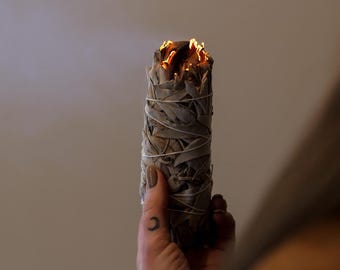 Könnte beinhalten: Ein zylindrisches Bündel aus getrockneten Salbeiblättern, das mit einer dünnen weißen Schnur umwickelt ist. Die Oberseite des Bündels brennt mit kleinen orangefarbenen Flammen und glühenden Funken und stößt eine feine Rauchfahne aus.