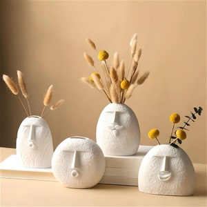 May include: Four white ceramic vases shaped like faces with different expressions. The vases are decorated with raised lines and have a textured surface. The vases are filled with dried flowers.