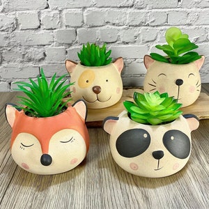 May include: Four ceramic planters shaped like animal faces, each with a small succulent plant. The planters are painted in neutral colours with black details. The animals are a fox, a cat, a dog, and a panda.