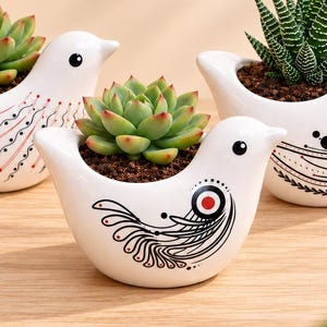 May include: Three white ceramic bird-shaped planters, each containing a small succulent plant. The planters feature black and red line art designs on their sides, including swirls, dots, and geometric patterns, with dark soil visible.