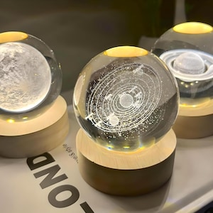 May include: Three glass globes with wooden bases, each containing a different celestial design. The globes are illuminated from within, creating a warm glow. The designs include a moon, a solar system, and a planet with rings.