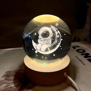 May include: A glass globe night light with a wooden base. The globe features an astronaut sitting on a crescent moon with stars in the background.