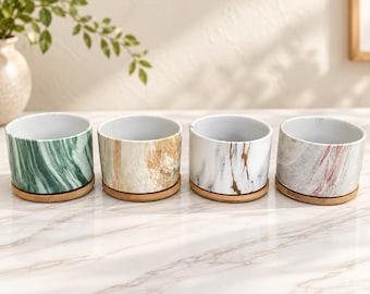 Op de afbeelding: Vier cilindervormige keramische plantenbakken met marmer-effect patronen in groen, beige, wit met gouden aders en grijs met roze strepen. Elke plantenbak staat op een ronde bamboe schotel en heeft een gladde, witte binnenkant.
