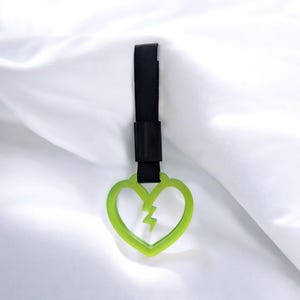May include: A neon green heart-shaped charm with a lightning bolt design, suspended by a black ribbon. The charm is made of a translucent material and hangs against a white background.