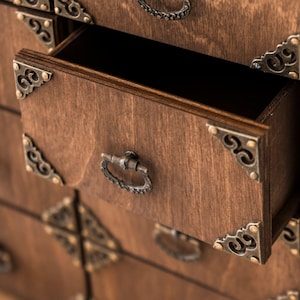 Könnte beinhalten: Eine Holzkiste mit mehreren Schubladen, jede mit einem dekorativen Metallgriff und Eckverzierungen. Die Schubladen sind geöffnet und zeigen das Innere der Kiste.