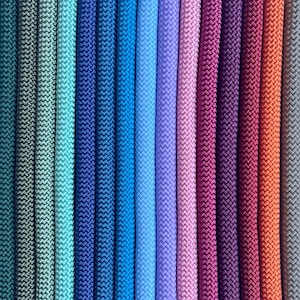 May include: A collection of vertical braided cords arranged in a gradient pattern, transitioning through shades of teal, blue, purple, magenta, and orange. Each cord features a consistent, tight, woven texture running in parallel lines.