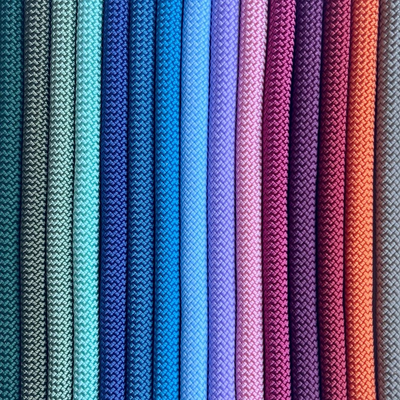 May include: A collection of vertical braided cords arranged in a gradient pattern, transitioning through shades of teal, blue, purple, magenta, and orange. Each cord features a consistent, tight, woven texture running in parallel lines.