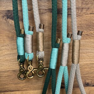 May include: A collection of six braided nylon cords in shades of dark green, light mint, and beige. Each cord features metallic end caps and decorative thread wrapping. Three cords are attached to brass swivel snap hooks with a distinct hook design.